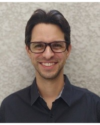 Homem de cabelos pretos, óculos de grau preto, sorrindo, com camisa preta