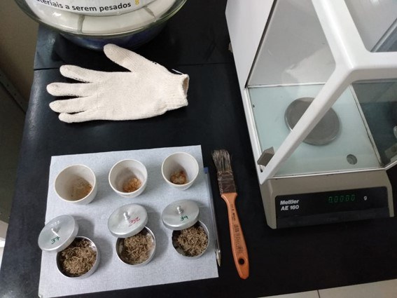 Imagem com pincel para limpeza, cadinhos e copos para análises, luva na cor branca, uma balança de precisão e matérias sobre uma mesa