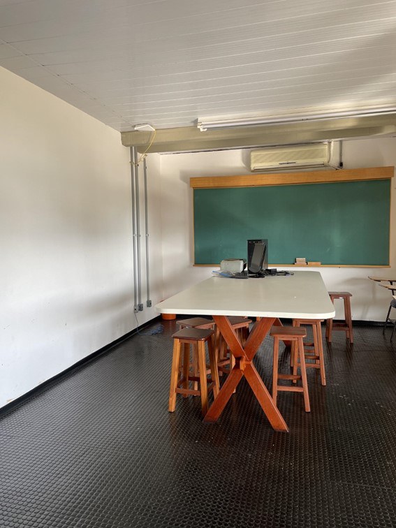 Foto interna do Laboratório de Análise Sensorial, com piso emborrachado preto, contendo uma mesa com pés de madeira e tampo branco, bancos de madeira em volta da mesa. Na mesa está um computador. Atrás da mesa estão uma lousa verde e um ar-condicionado